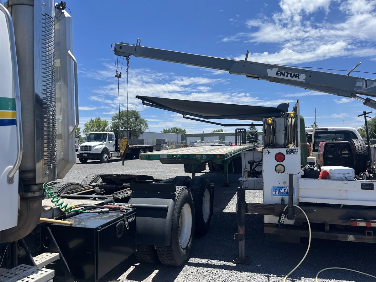A flatbed truck with a boom crane lifting a deck section beside a semi tractor in the yard.