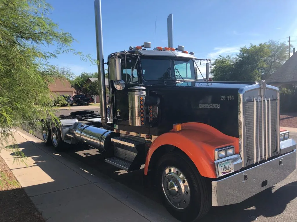 An orange and black Kenworth W900 day cab parked in the sun outside the shop.
